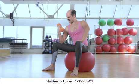 Full Height. Bald Funny Man In A Pink T-shirt Is Training With Dumbbells On A Sports Ball In The Gym.