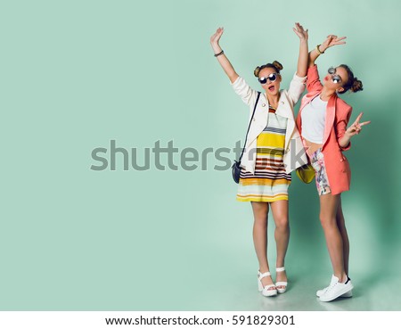 Full height image  of two happy cheeky girls  , best friends having fun , laughing on blue background. . Wearing stylish  casual jackets, striped pop dress. Space for text. 