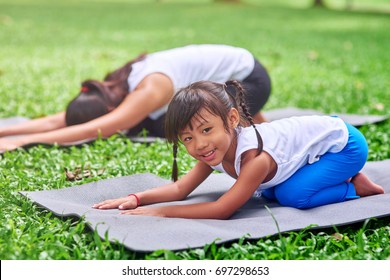 Full of joy. Positive cute little thai sitting on the roll mat and resting in the park while doing stretching exercises with her mother - Powered by Shutterstock