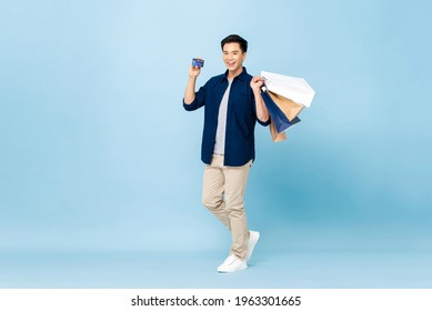 Full Lenght Portrait Of Happy Smiling Handsome Asian Man Carrying Shopping Bags And Showing Credit Card In Light Blue Isolated Studio Background