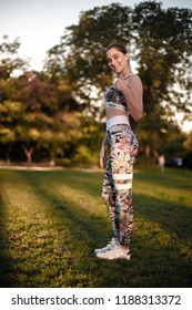 Full Lenght Side View Portrait Of A Healthy Brunette Sports Fitness Slim Woman In Top With Tropical Print Outdoors In The Park On Green Grass With Sundown Light Looking Camera Showing Thumbs Up.