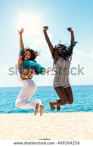Full length action portrait of black and white women laughing and jumping together on beach.