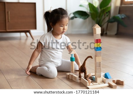 Full length adorable interested small asian vietnamese ethnicity preschool baby girl sitting on warm wooden floor, playing with colorful wooden cubes constructing building alone in living room.