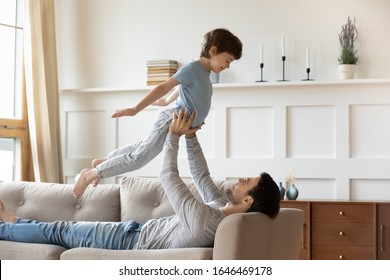 Full length affectionate young man lying on cozy sofa, lifting little schoolboy child. Joyful father practicing acroyoga with small son in living room. Two generations family playing together indoors.