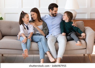 Full length affectionate young parents relaxing on sofa, cuddling small children. Happy couple enjoying free leisure weekend time together in living room, communicating talking chatting with kids.