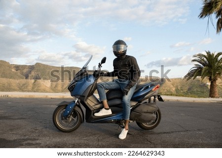 Full length of an African American biker sitting on his scooter. Arid and tropical background