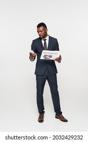 full length of african american businessman in suit using smartphone while holding newspaper isolated on grey