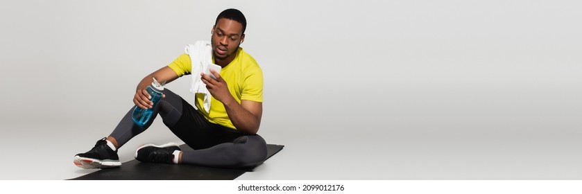 full length of african american sportsman sitting on fitness mat and using smartphone near sports bottle on grey, banner - Powered by Shutterstock