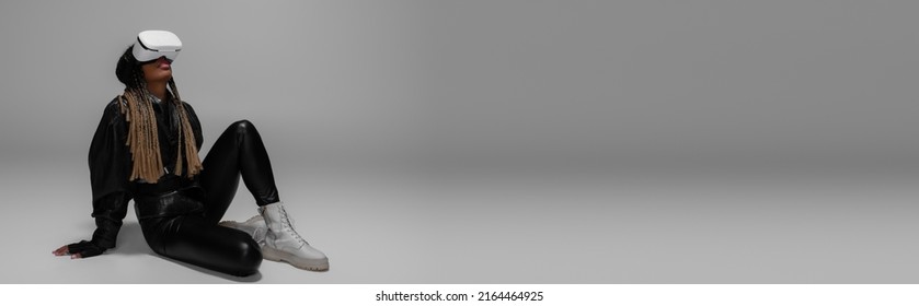 Full length of african american woman in vr headset and leather jacket sitting on grey background, banner