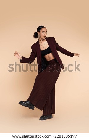 full length of african american woman in trendy maroon suit with wide leg pants posing on beige 商業照片 © 