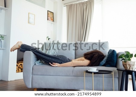 Full length of african girl lying face down on sofa near table with coffee cup, feeling tired after sleepless night or party. Exhausted sleepy black woman in dress and boots sleeping on couch at home