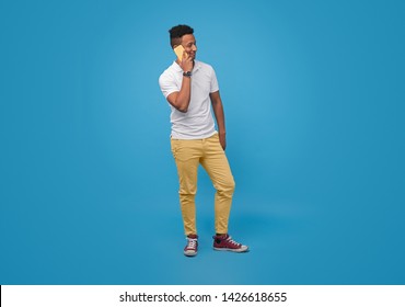 Full Length African Guy In Casual Outfit Smiling And Looking Away While Answering Phone Call Against Blue Background