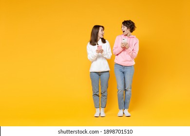 Full length of amazed two young women friends 20s in casual white pink hoodies using mobile cell phone typing sms message looking at each other isolated on bright yellow background studio portrait