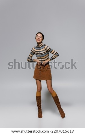 full length of amazed young woman in striped turtleneck with skirt and boots posing on grey