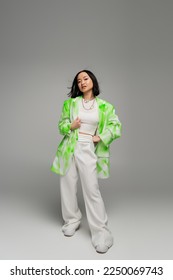 full length of asian woman in trendy oversize outfit posing with hand on hip on grey background