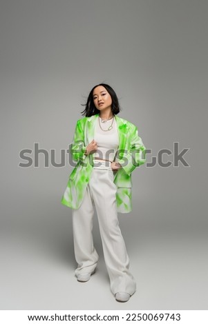 full length of asian woman in trendy oversize outfit posing with hand on hip on grey background