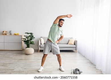 Full length of athletic young Arab man bending aside, doing exercises at home, copy space. Millennial Eastern man having domestic training, working out indoors during covid lockdown