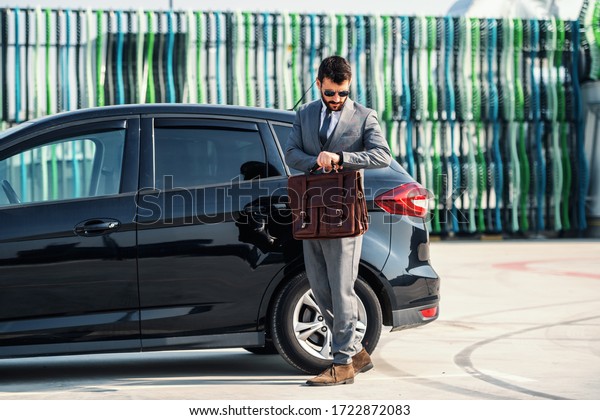 Full length of\
attractive caucasian rich businessman standing next to his\
expensive car, holding briefcase and looking at wristwatch. He\
don\'t want to be late for\
meeting.