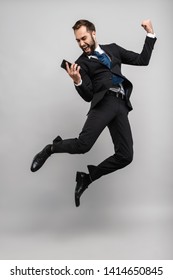 Full length of an attractive smiling young businessman wearing suit jumping isolated over gray background, wearing earphones, holding mobile phone