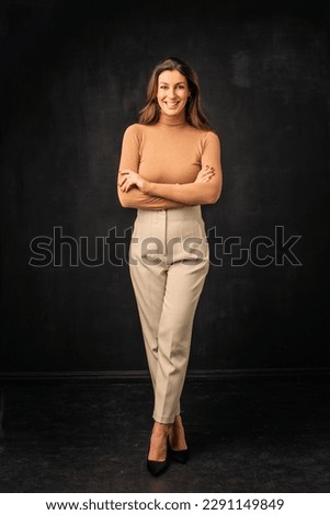 Full length of an attractive woman toothy smiling and looking at camera. Brunette haired female wearing turtleneck sweater and trousers while standing against isolated black background. Copy space. 