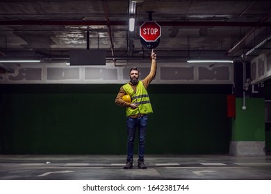 Full Length Of Attractive Worker In Vest, With Helmet Under Armpit Standing Inside Of Underground Parking Lot In Construction Process And Pointing At Stop Sign On Ceiling.