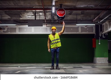 Full Length Of Attractive Worker In Vest, With Helmet On Head Standing Inside Of Underground Parking Lot In Construction Process And Pointing At Stop Sign On Ceiling.