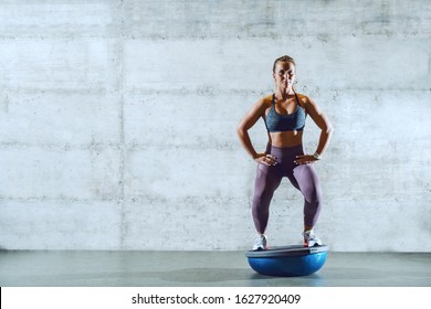 Full Length Of Attractive Young Caucasian Sportswoman In Sportswear With Ponytail Standing On Bosu Ball And Doing Squat Endurance,