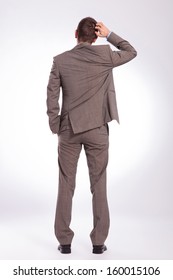 Full Length Back View Picture Of A Young Business Man Scratching His Head While Holding A Hand In His Pocket. On A Gray Background