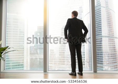 Full length back view of successful businessman in suit standing in office with hands on his waist, CEO looking through window at big city buildings, planning new project, waiting for meeting to start