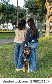 A full length back view of two sisters or friends standing together on green grass with a loyal companion dog wearing a blue harness. Shot in a public park surrounded by trees