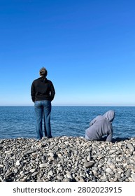 Full length back view of two people relaxing on a stony sea shore, enjoying a calm sunny day with a massive clear blue sky background and plenty of copy space for text.