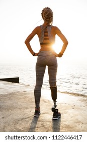 Full Length Back View Of Young Disabled Athlete Woman With Prosthetic Leg Doing Exercises While Standing At The Beach