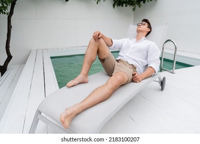 Full Length Of Barefoot Man In Summer Clothes Resting On Deck Chair At Poolside