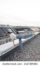 Full Length Of Barefoot Woman In Jeans Looking Down Rooftop Of Urban Building