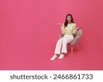 Full length beautiful Asian teen woman using smartphone and sitting on chair isolated on pink copy space background. People lifestyle Technology Concept.
