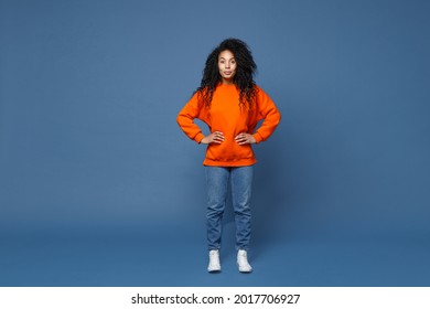 Full length of beautiful attractive young african american woman in casual bright orange sweatshirt standing with arms akimbo on waist looking camera isolated on blue color background studio portrait