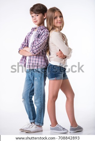 Full length of beautiful little girl and boy looking at camera and smiling while standing with crossed arms back to back on light background