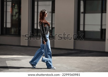 Full length beautiful young brunette woman Wearing black leather jacket and wide blue jeans, sneakers, sunglasses and small handbag with chain, walking street on sunny day. 商業照片 © 
