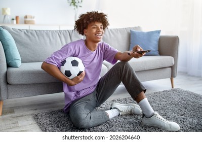 Full Length Of Black Teen Guy Watching Football On TV, Using Remote Control, Holding Soccer Ball At Home. Funky Afro Teenager Supporting Favorite Team, Sitting On Floor, Following Sports Championship