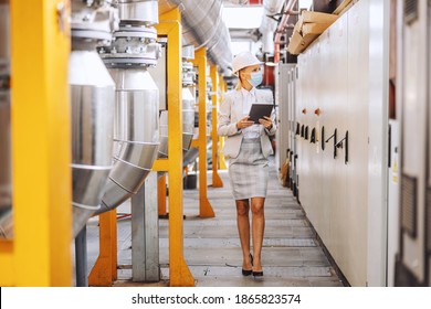 Full Length Of Blond Female Boss In Formal Wear, With Helmet On Head, With Protective Mask Walking Trough Heating Plant And Using Tablet To Check On Machinery.