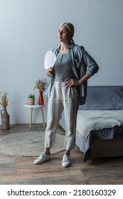 Full Length Of Blonde Woman With Menopause Suffering From Heat And Holding Fan In Bedroom