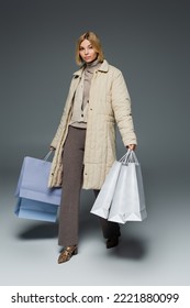Full Length Of Blonde Young Woman In Puffer Coat Holding Shopping Bags While Walking On Grey