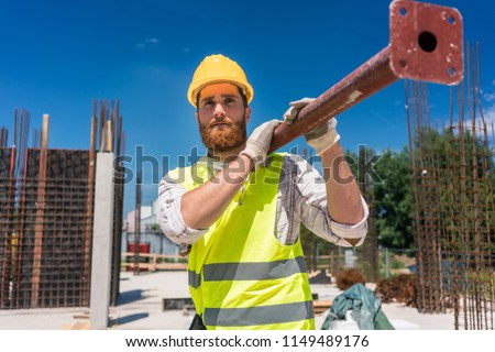Full length of a blue-collar worker, wearing safety equipment while carrying a heavy metallic bar during work on the construction site of a residential building