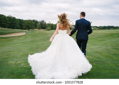 Full Length Body Portrait Of Young Bride And Groom Running On Green Grass Of Golf Course, Back View. Happy Wedding Couple Walking Through Golf Course, Copy Space