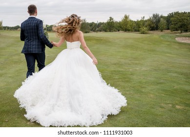 Full Length Body Portrait Of Young Bride And Groom Running On Green Grass Of Golf Course, Back View. Happy Wedding Couple Walking Through Golf Course
