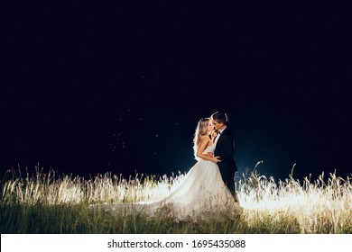 Full Length Body Portrait Of Young Bride And Groom Enjoying Romantic Moments Outside In The Night. Wedding Couple. Standing Face To Face With Backlight.