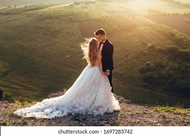 Full length body portrait of young bride and groom enjoying romantic moments outside at sunset in beautiful summer day. Wedding couple. Standing face to face with the green hills on background.