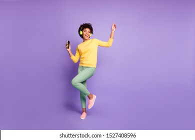 Täyspitkä kehon koko kuva trendikäs tyylikäs söpö vapaa tyttöystävä yllään vihreät housut housut keltainen villapaita jalkineet kuulokkeissa musiikin kuuntelu tanssia eristetty violetti pastelliväri tausta