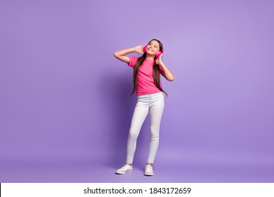 Full Length Body Size Photo Of Dreamy Little Girl Smiling Listening To Favourite Songs With Headphones Wearing T-shirt Jeans Isolated On Purple Color Background