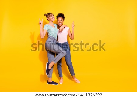 Full length body size photo beautiful she her lady best buddies different nationalities hold hands arms v-sign say hi hello wear casual white striped t-shirt clothes outfit isolated yellow background